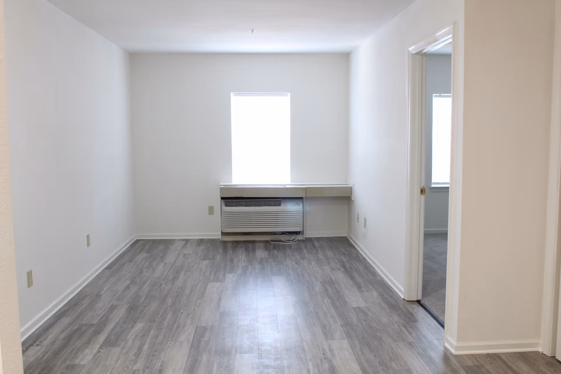 Empty bright room with gray wood-look flooring, a central window above a wall air conditioner and a doorway to an adjacent room.