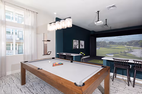 Bright interior recreation room with a pool table in the foreground and a golf simulator screen and seating area in the background.