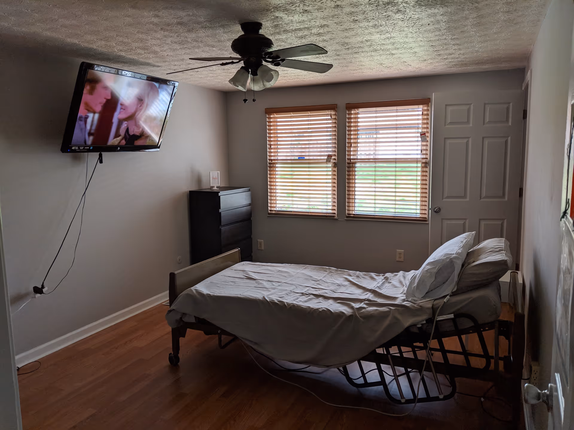 A simple bedroom in a senior care home with a hospital-style adjustable bed, two pillows, a wooden dresser, a wall-mounted TV showing a scene with two people, two windows with wooden blinds, a ceiling fan with lights, and a closed door.
