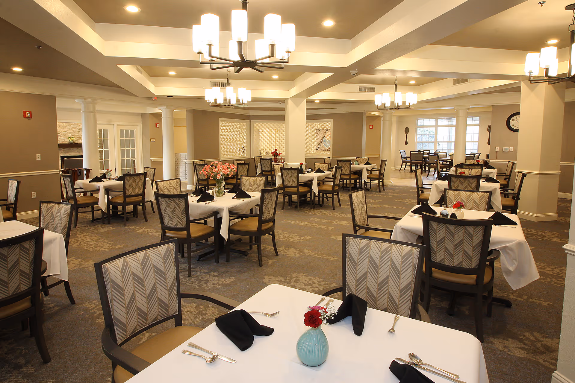 A spacious dining room in a senior living facility with multiple tables covered in white tablecloths, each set with black napkins, silverware, and small flower vases. The room features patterned carpet, beige walls, white columns, and modern chandeliers hanging from the ceiling. Large windows allow natural light to enter the room.