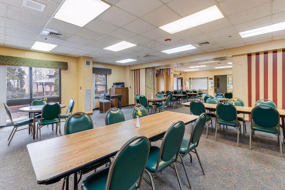 A spacious dining room with multiple rectangular and round tables surrounded by green cushioned chairs. The room has large windows with views of an outdoor gazebo and trees. The walls are painted yellow with one wall featuring red vertical stripes. The ceiling has fluorescent lighting panels, and there are small flower vases on some tables.