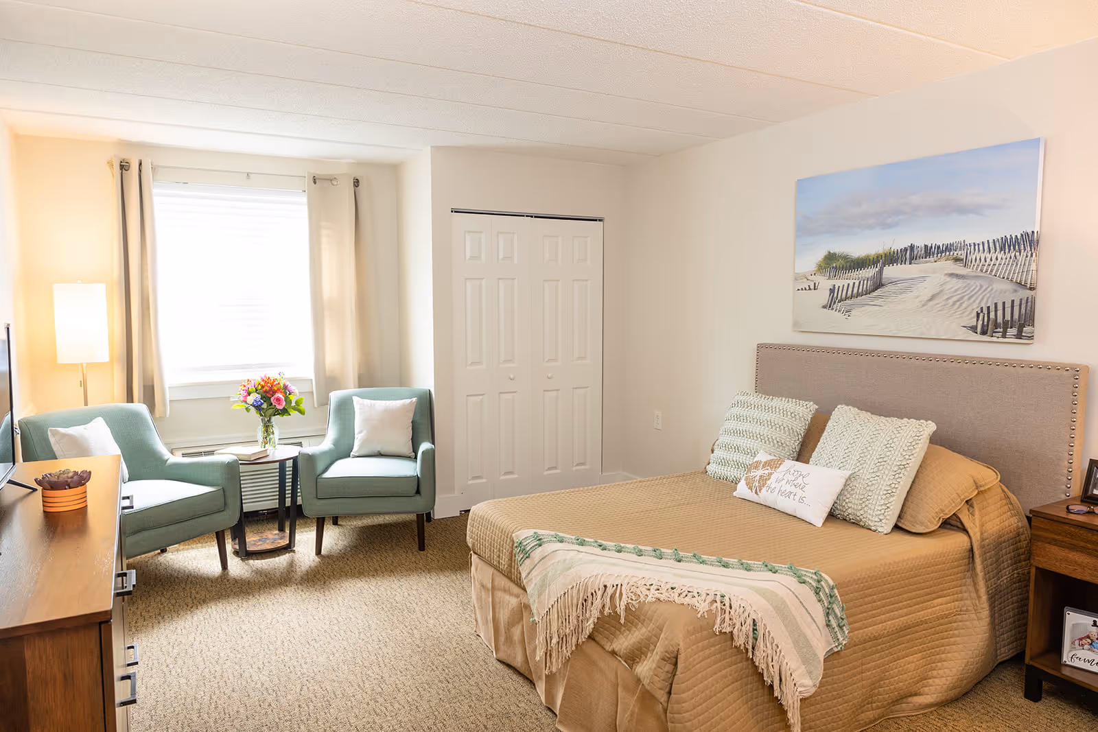 A well-lit bedroom in a senior living facility featuring a neatly made bed with beige bedding and decorative pillows, two light blue armchairs with a small table and a vase of flowers between them near a window with beige curtains, a wooden dresser with a small plant, a floor lamp, and a large beach-themed painting on the wall above the bed.