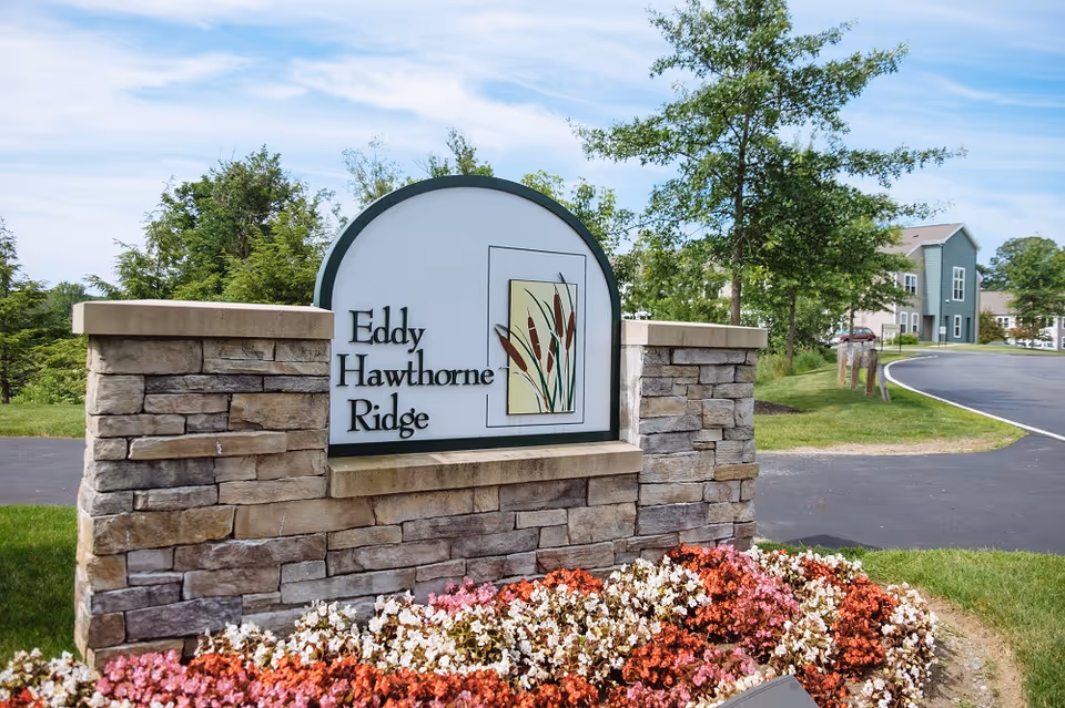 Stone entrance sign reading "Eddy Hawthorne Ridge" surrounded by flowers and landscaping with residential buildings and a driveway in the background.