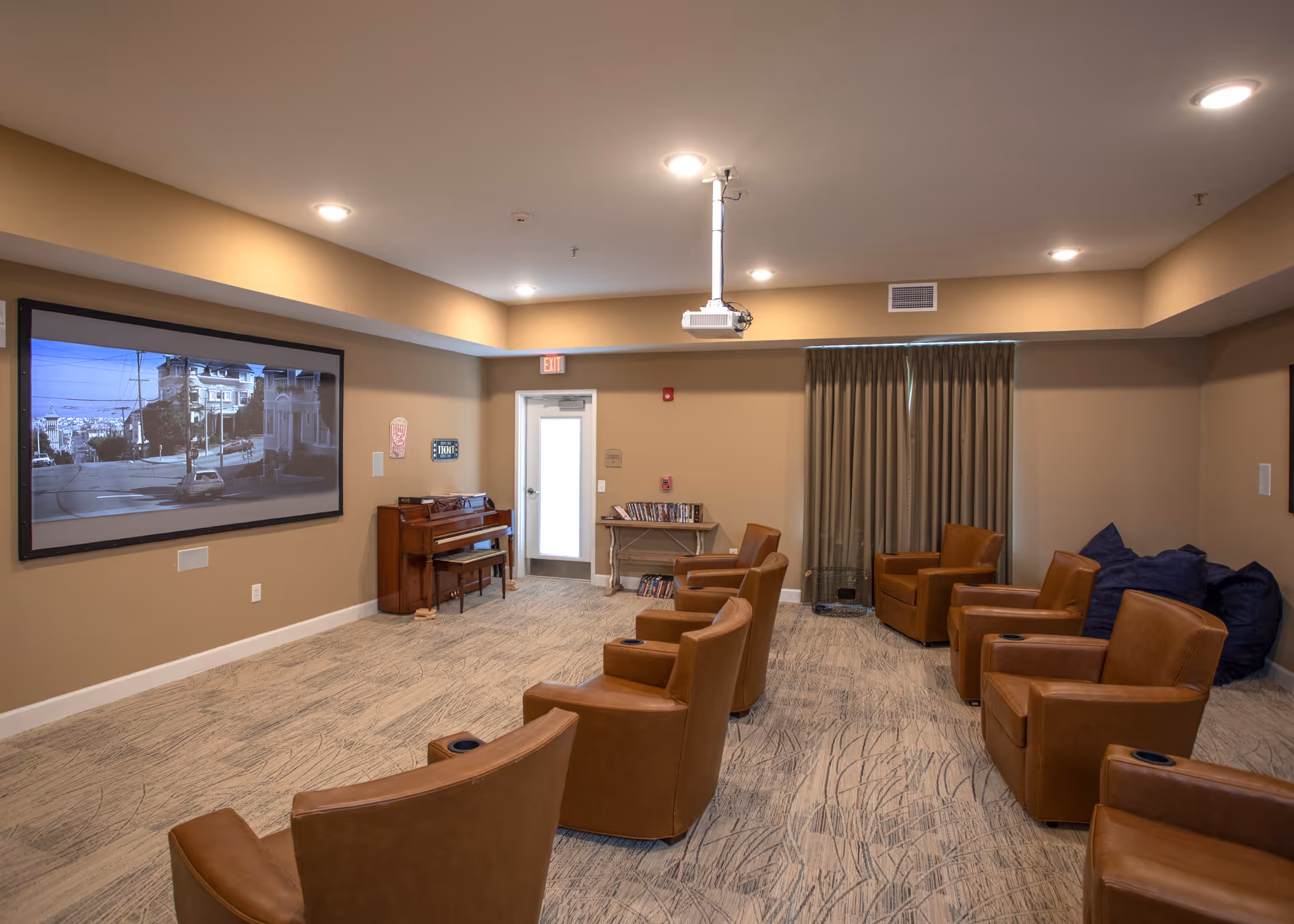 A cozy media room with several brown leather armchairs arranged in rows facing a large wall-mounted screen displaying a street scene. The room has beige walls, carpeted floor, a piano against one wall, a small table with DVDs, and a ceiling-mounted projector. There are curtains covering a window and an exit door at the back.
