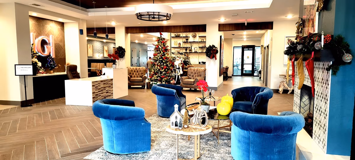 A decorated senior living lobby with blue velvet chairs around tables, a Christmas tree, reception desk, and a fireplace with stockings.