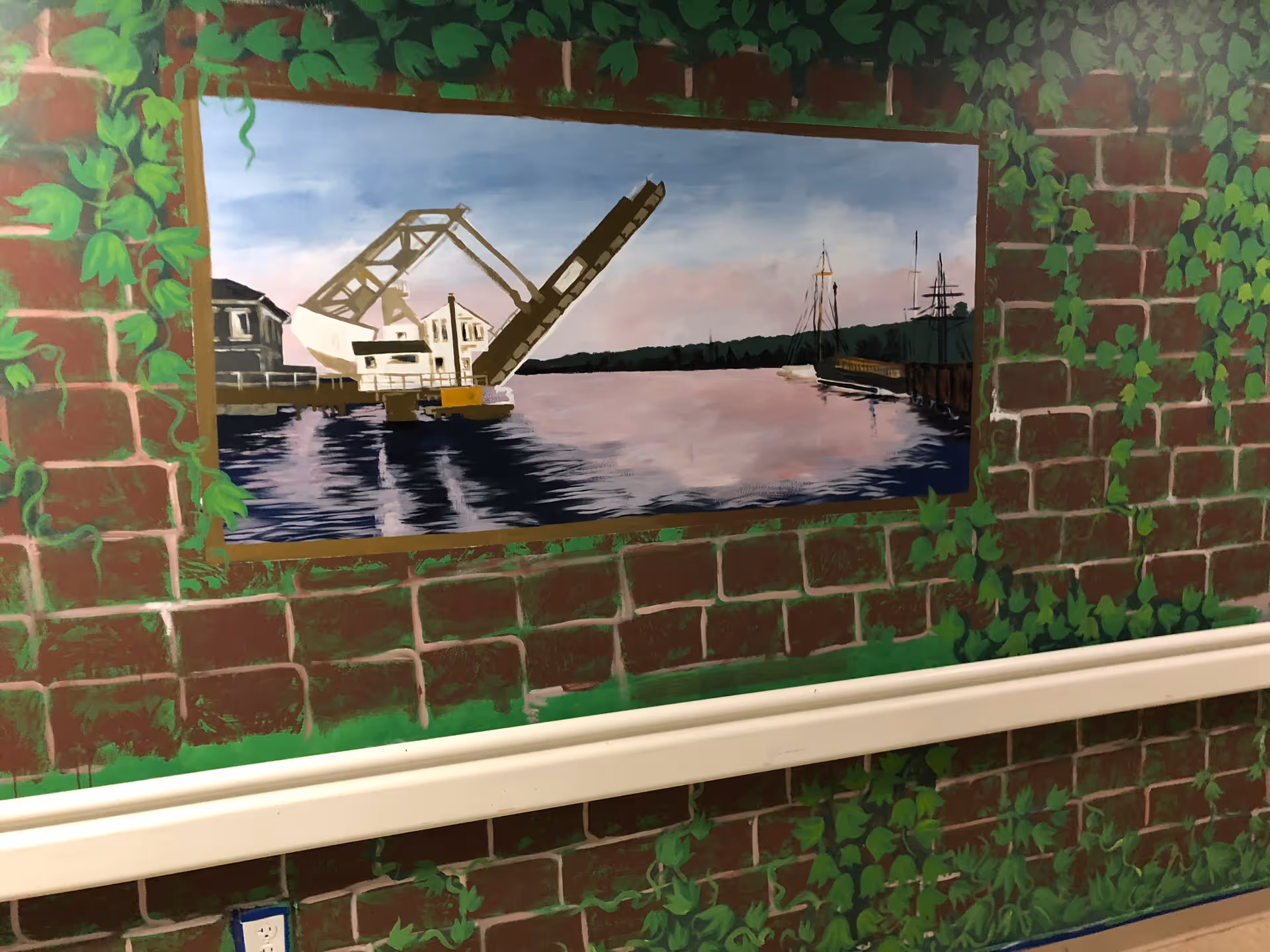 A mural painted on a brick wall with green ivy. The mural depicts a waterfront scene with a raised drawbridge, buildings, and boats on calm water under a blue and pink sky.