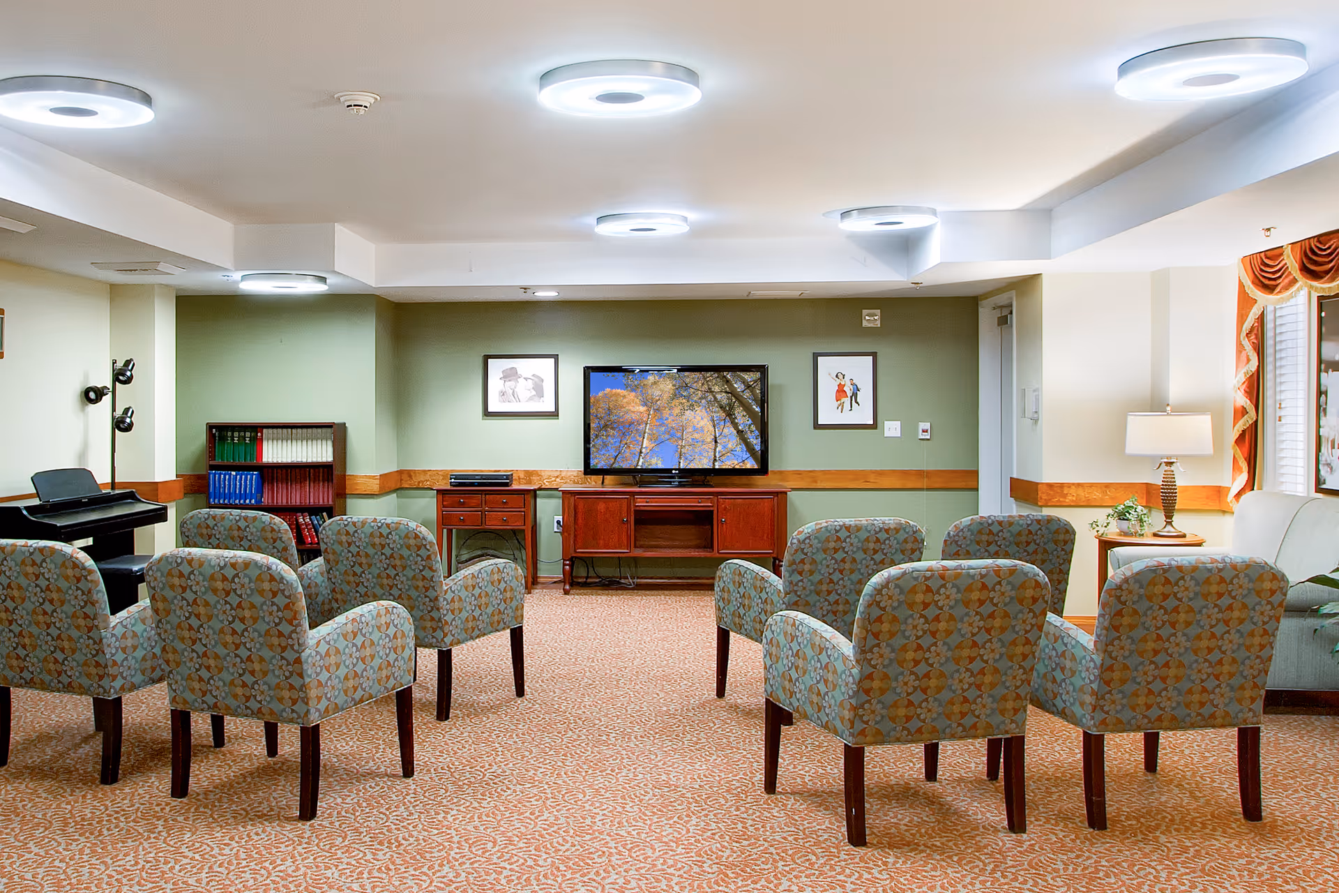A cozy living room area with eight patterned armchairs arranged in two rows facing a flat-screen TV mounted on a green wall. The TV displays an image of tree branches with autumn leaves against a blue sky. The room has a patterned carpet, modern circular ceiling lights, a piano with a bench on the left, a bookshelf with colorful books, framed pictures on the wall, a side table with a lamp, and a window with orange curtains on the right.