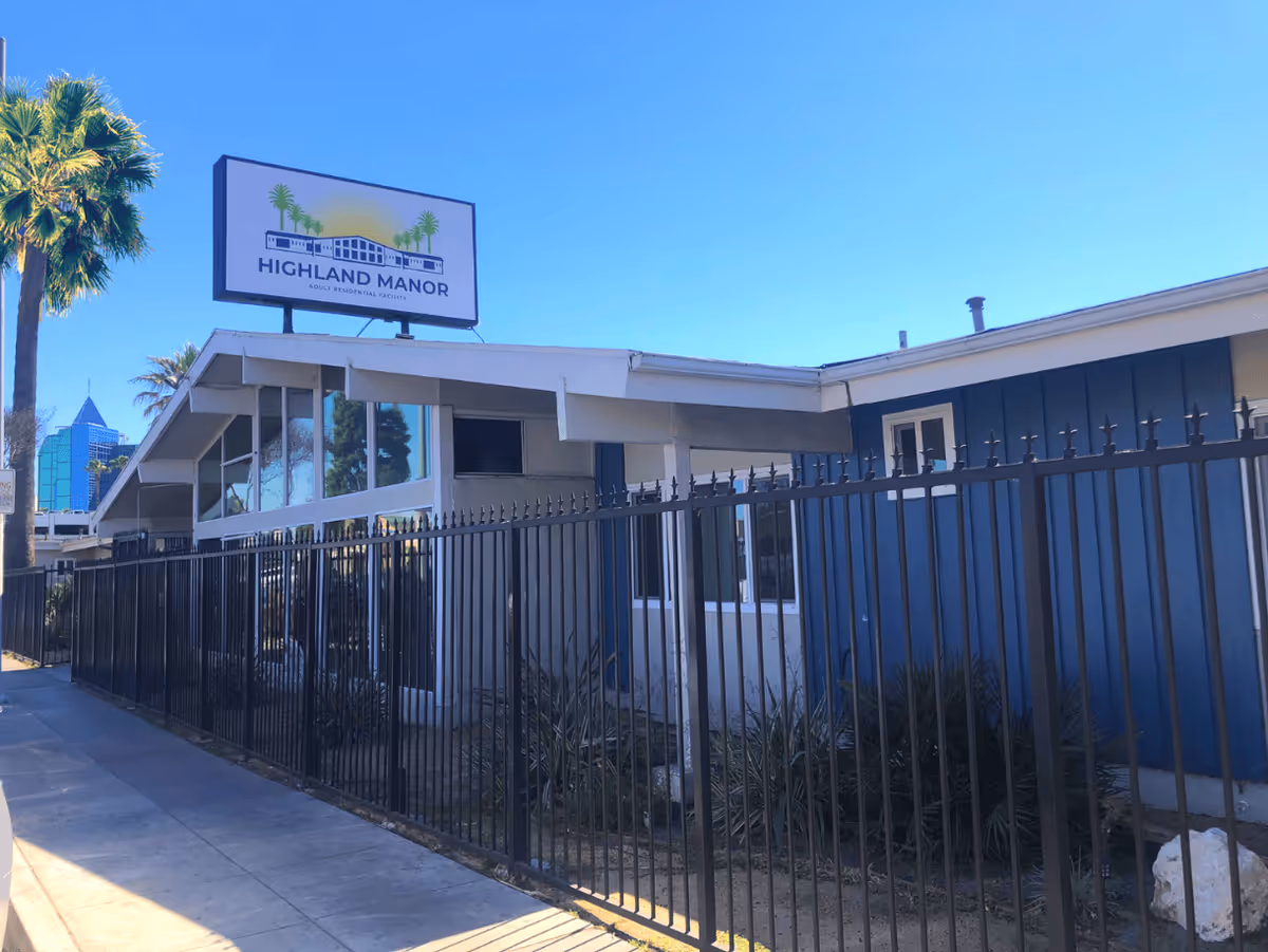 Exterior view of Highland Manor Assisted Living facility with a black metal fence in front, a sidewalk, palm trees, and a clear blue sky.