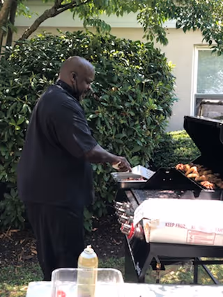 A man wearing a black shirt is grilling food outdoors on a barbecue grill. The grill is open, showing food cooking inside. There are green bushes and a tree in the background, along with part of a building with a window.