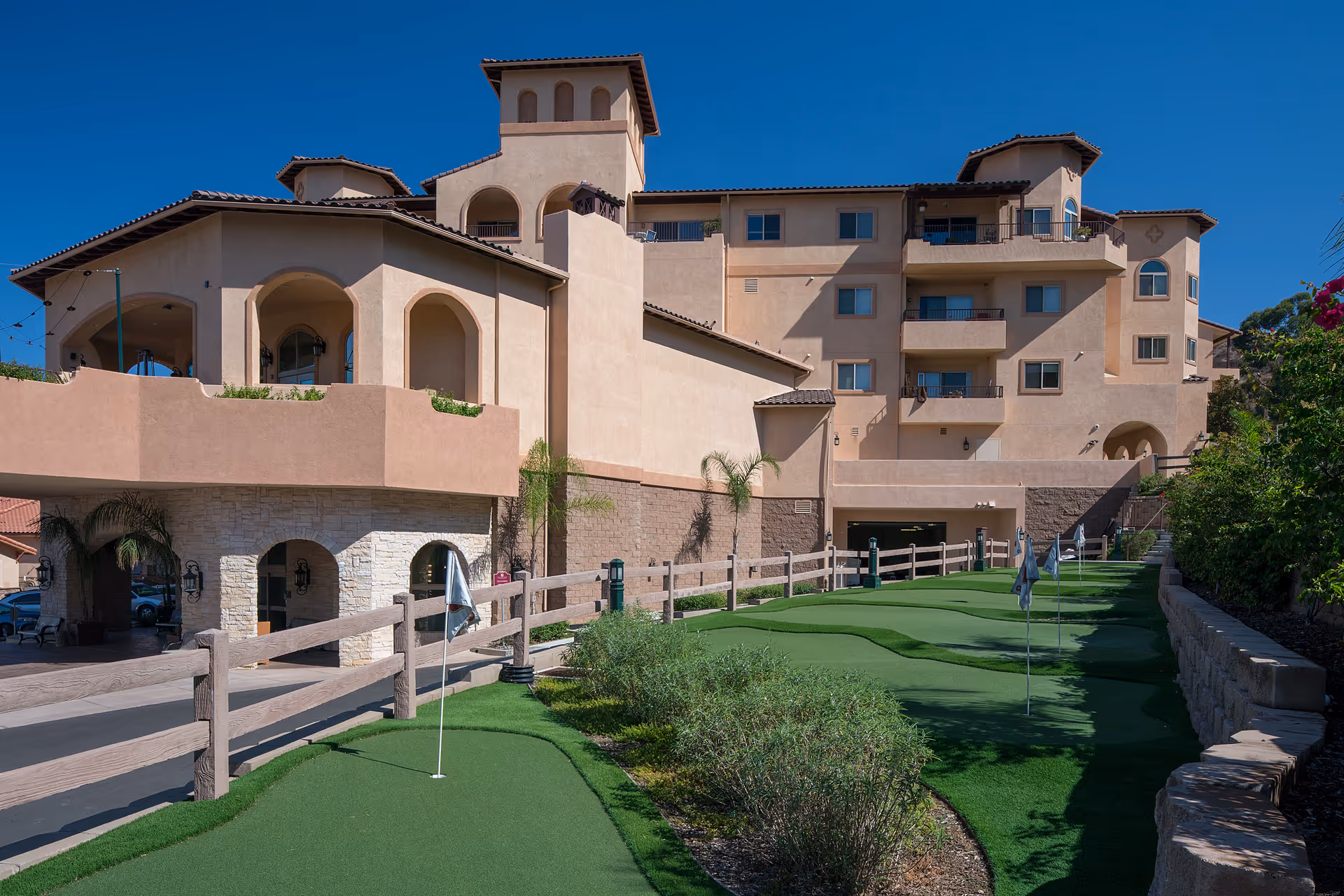 Exterior view of a multi-story senior living facility with beige stucco walls and a tiled roof under a clear blue sky. In the foreground, there is a putting green with several golf flags and a wooden fence along the side.