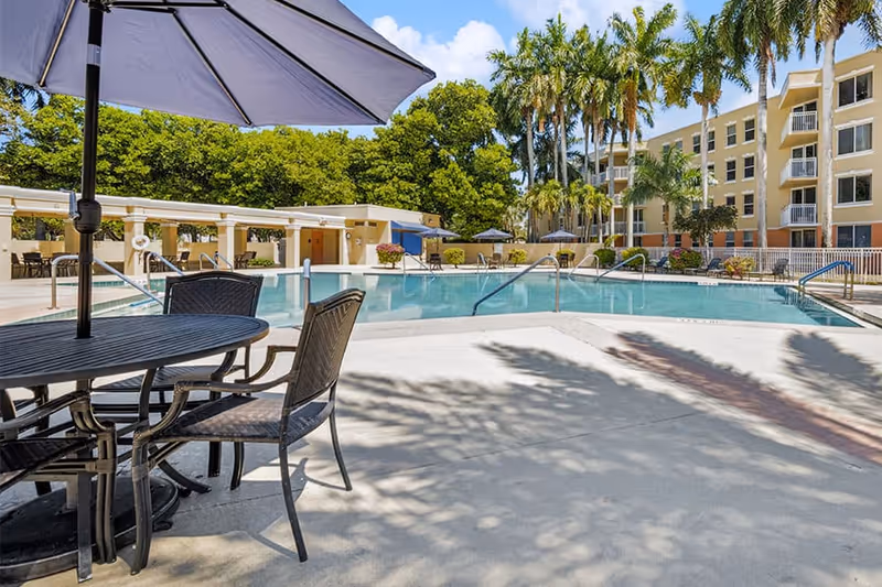 Outdoor swimming pool area with metal table and chairs under a large umbrella in the foreground. Palm trees and a multi-story residential building are visible in the background under a blue sky with some clouds.