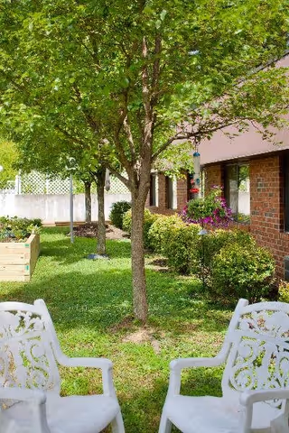 Outdoor garden area with green grass, a tree in the center, bushes, and flowers near a brick building. Two white decorative chairs are positioned facing each other in the foreground.