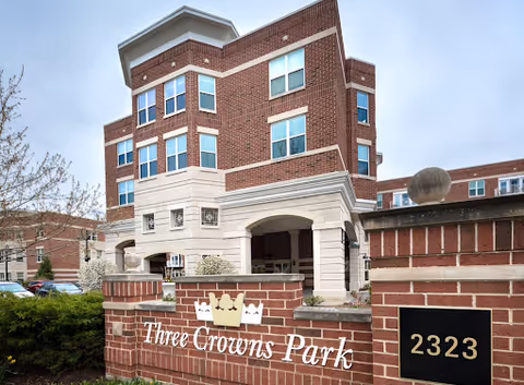 Brick multi-story senior living building entrance with a 'Three Crowns Park' sign and address plaque reading 2323.