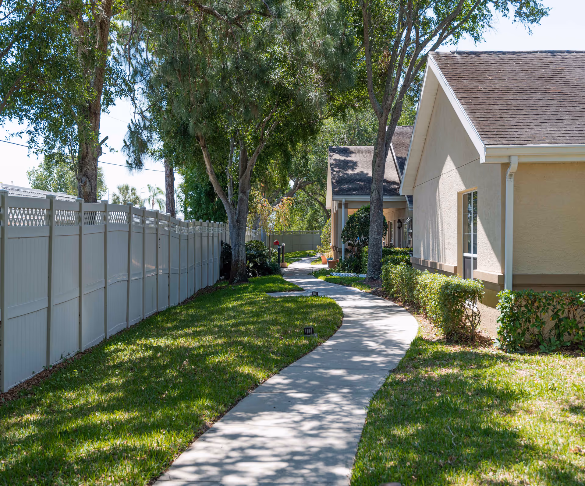 A winding concrete pathway bordered by green grass and trees leads alongside a beige building with a shingled roof. A tall white fence runs parallel to the path on the left side, and there are small bushes and plants near the building on the right. The scene is outdoors on a sunny day with shadows cast by the trees.
