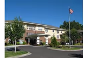 Front exterior of a three-story senior living building with a covered entrance, circular driveway, landscaping, and an American flag on a flagpole.