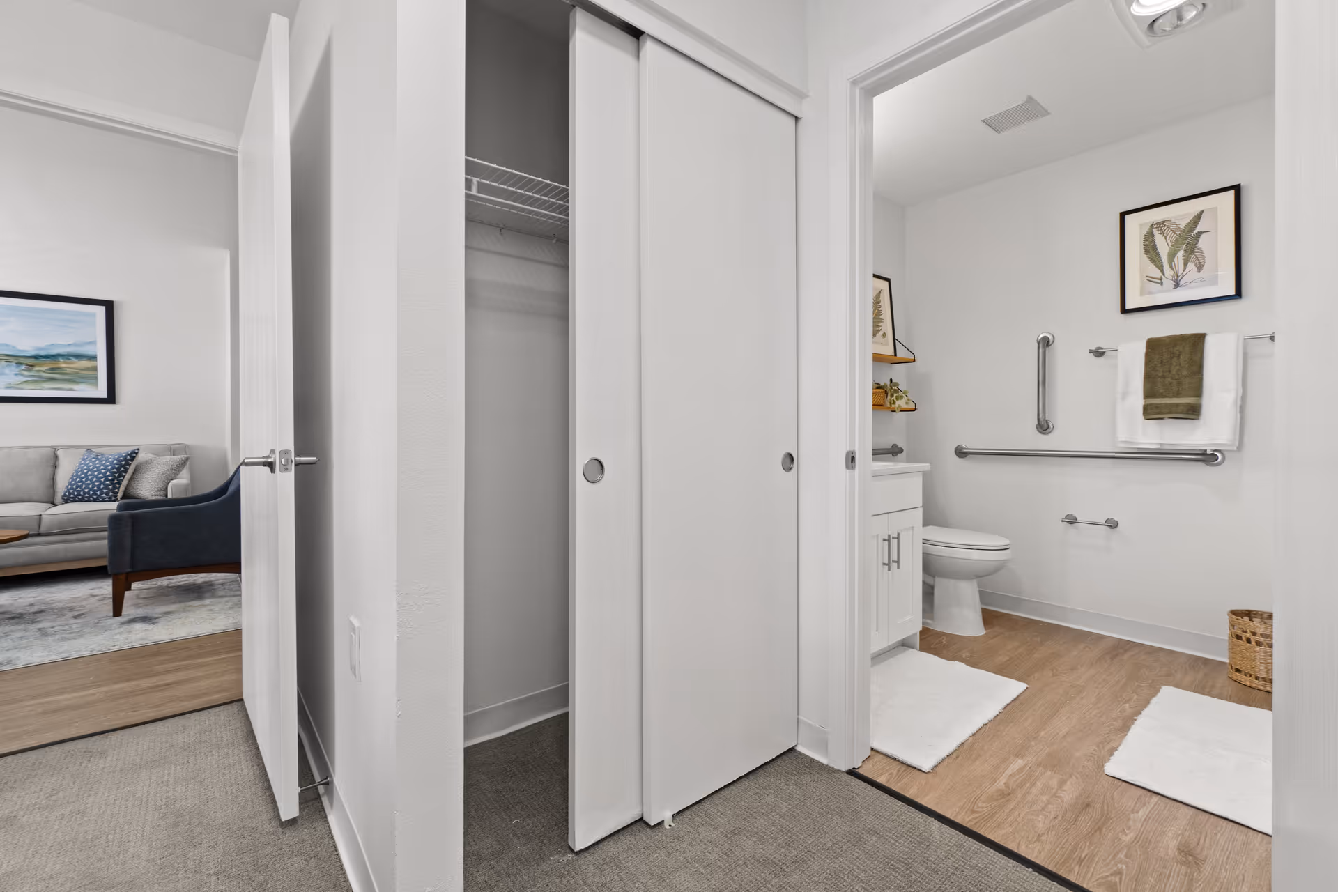 View of a hallway leading to a bathroom with white walls and wooden flooring. The bathroom features a toilet, grab bars, a towel rack with a green and white towel, a framed botanical print on the wall, and a white vanity with a mirror. To the left of the hallway is an open closet with sliding doors, and beyond that, a living room area with a gray sofa, a blue armchair, a coffee table, and a landscape painting on the wall.
