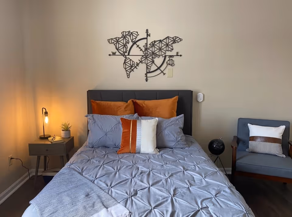 A neatly made bed with gray bedding and orange accent pillows in a bedroom with wall art above the headboard, a nightstand with a lamp and plant, and an armchair to the right.