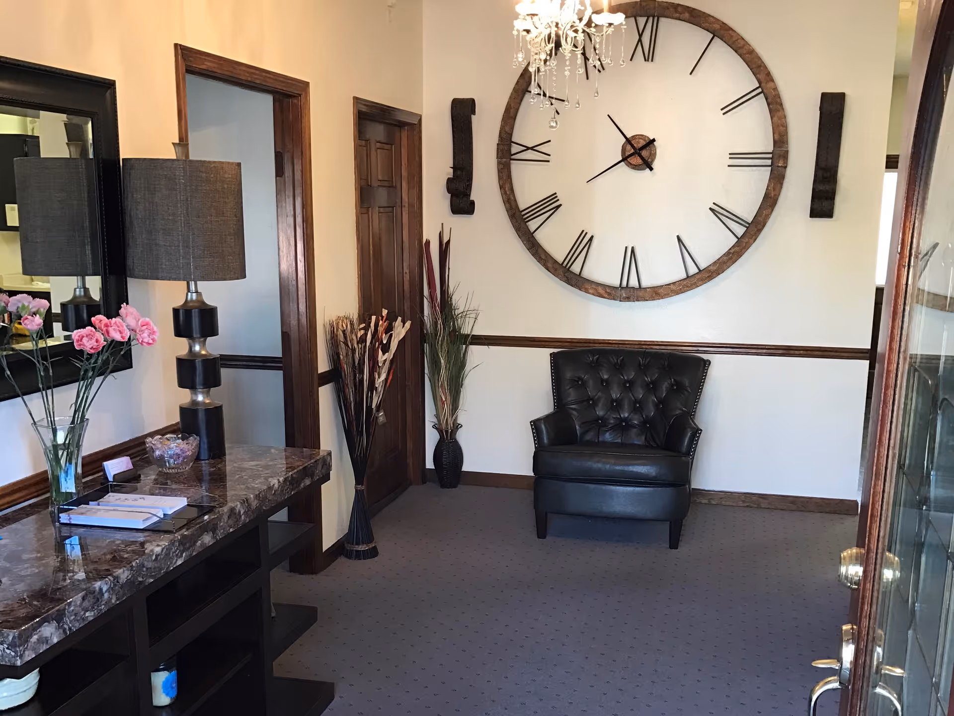 An interior room with a large wall clock featuring Roman numerals, a black leather armchair, a marble-top console table with a lamp, a vase of pink flowers, and decorative plants in vases. The room has beige walls with wooden trim and a carpeted floor.