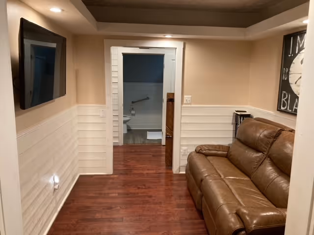 Interior view of a room with wooden flooring and beige walls. A brown leather couch is positioned on the right side, and a flat-screen TV is mounted on the left wall. Through an open doorway, a bathroom with a toilet and grab bar is visible.