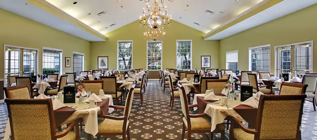 A spacious dining room with multiple tables covered in white tablecloths and brown overlays, each set with napkins, glasses, and menus. The room features green walls, large windows letting in natural light, and elegant chandeliers hanging from a high ceiling. Chairs with patterned upholstery surround the tables, and framed artwork is visible on the walls.