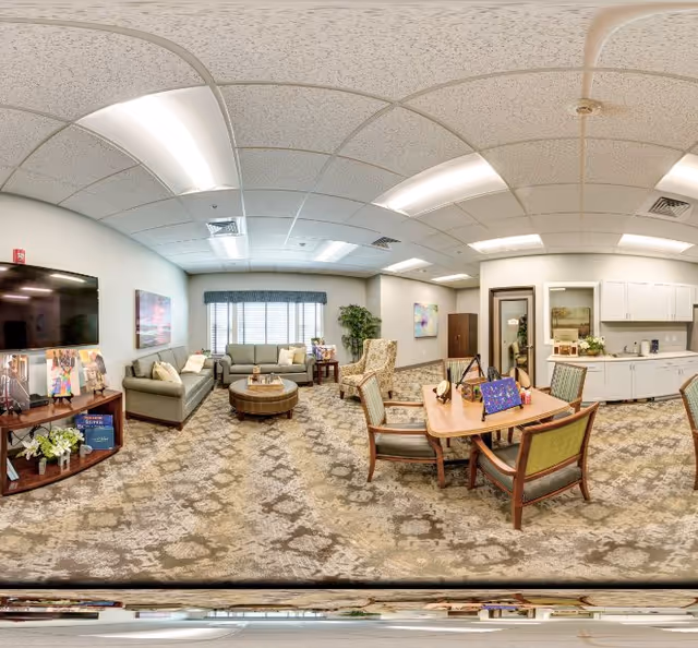 A bright communal lounge with sofas, armchairs, a round coffee table, a dining table, TV and kitchenette.