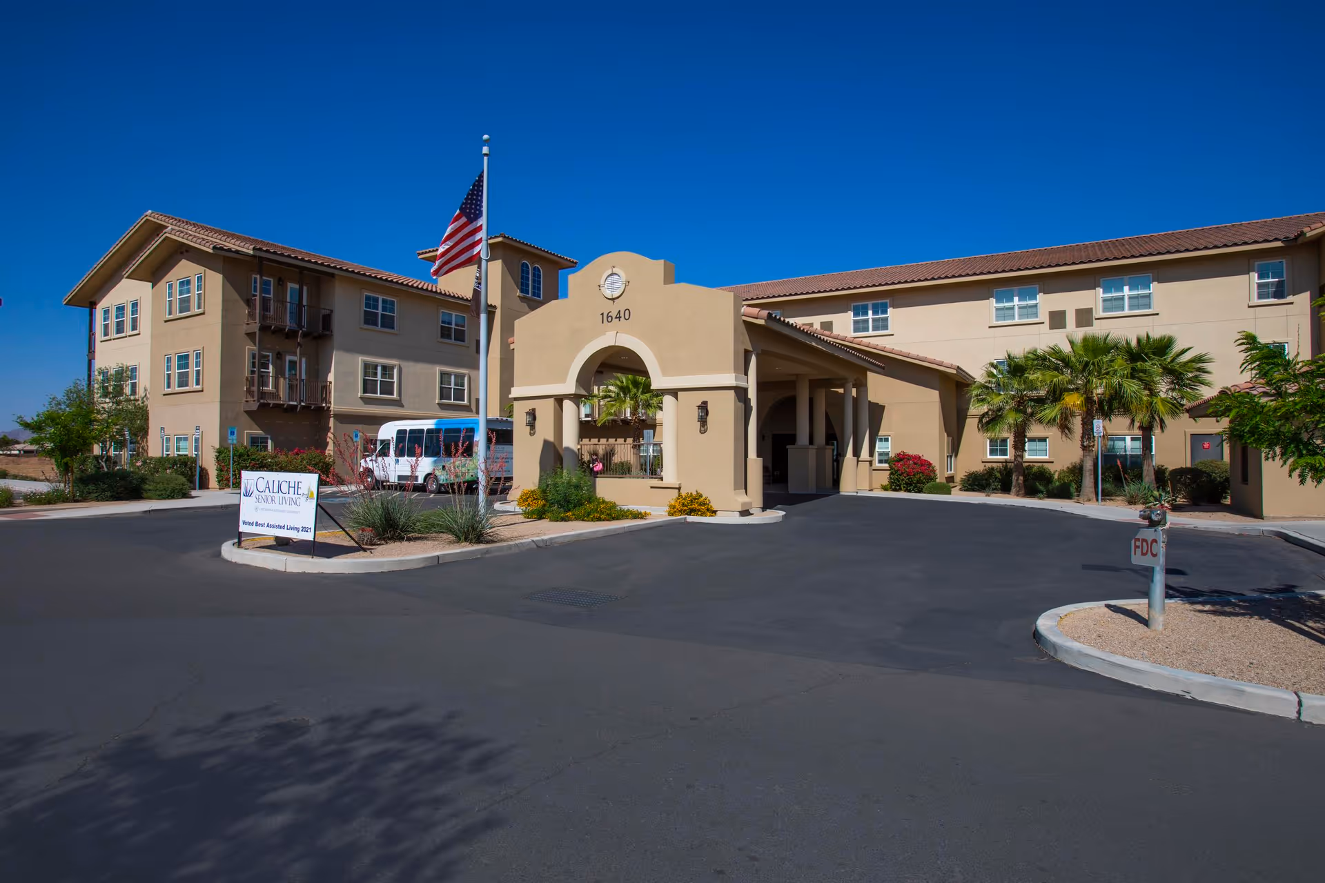 Exterior view of a senior living facility with a beige stucco building, an arched entrance with the number 1640, an American flag on a flagpole, palm trees, and a sign that reads 'Caliche Senior Living'.