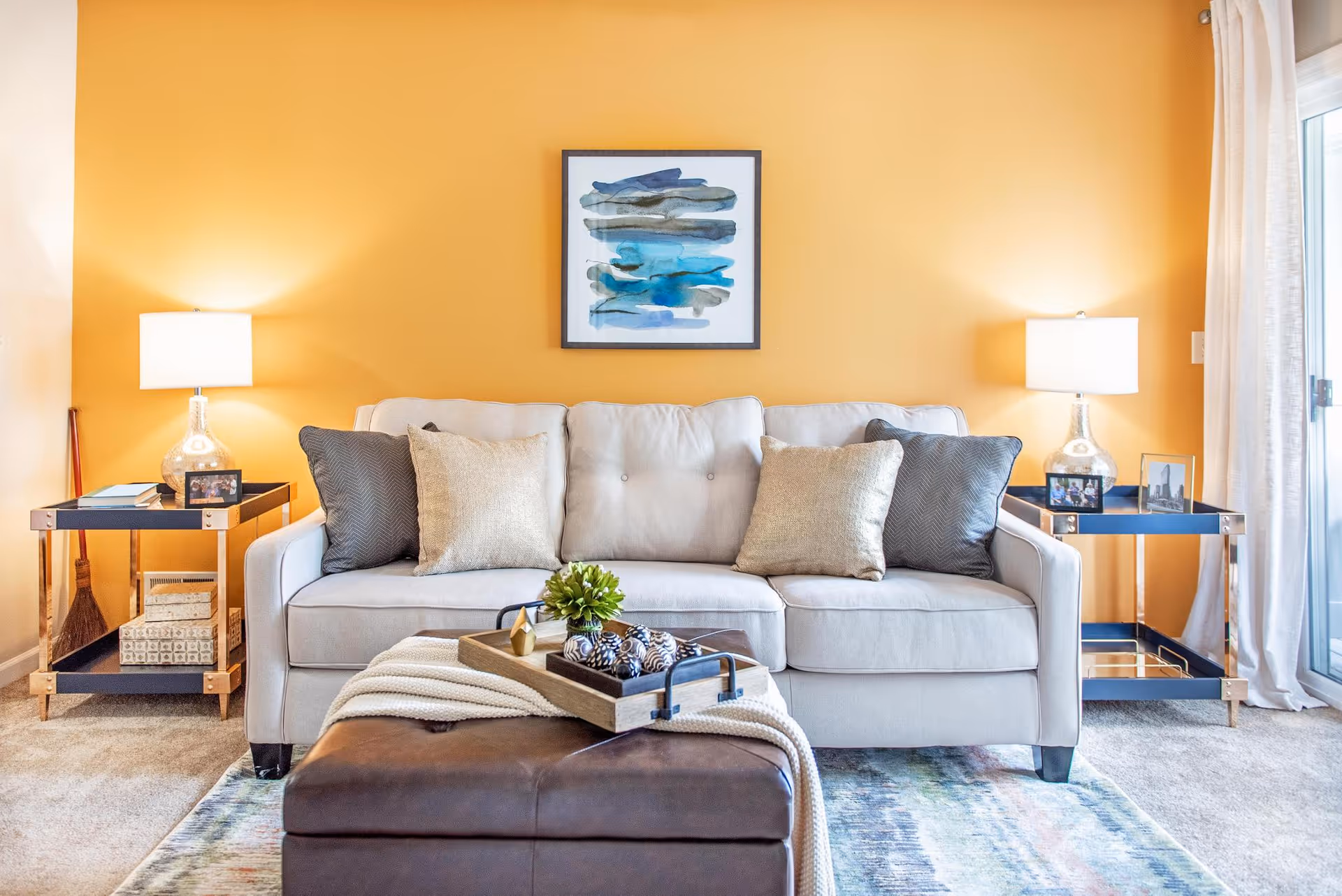 A cozy living room with a light beige sofa adorned with four cushions in neutral tones. On either side of the sofa are matching side tables with glass lamps, framed photos, and decorative items. A brown leather ottoman with a tray holding decorative objects and a blanket is placed in front of the sofa. The walls are painted a warm yellow, and a framed abstract painting with blue and gray tones hangs above the sofa. Light filters in from a window with white curtains on the right side.