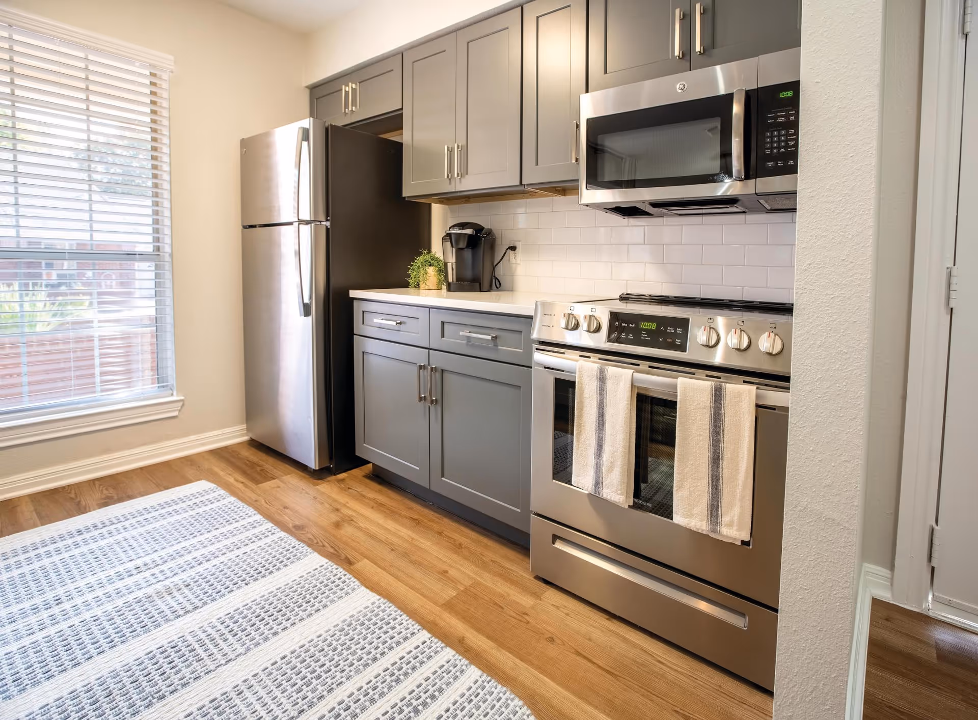 Modern kitchen with gray cabinets, stainless steel refrigerator, stove and microwave, white subway tile backsplash and a window with blinds.