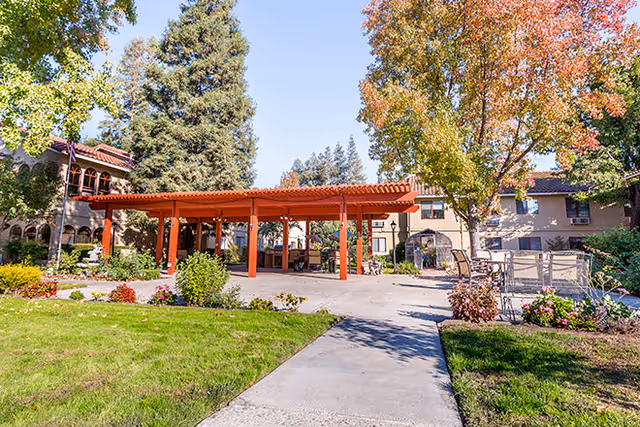 Outdoor courtyard area at Cogir of Manteca featuring a red pergola, concrete pathways, green grass, trees with autumn foliage, and outdoor seating with tables and chairs surrounded by flowers and shrubs.