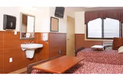Interior view of a senior living facility bedroom with two beds covered in red patterned bedspreads, a wooden coffee table in front, a wall-mounted sink with a mirror above it, a paper towel dispenser, a soap dispenser, a wall-mounted television, and a window with blinds and a valance.