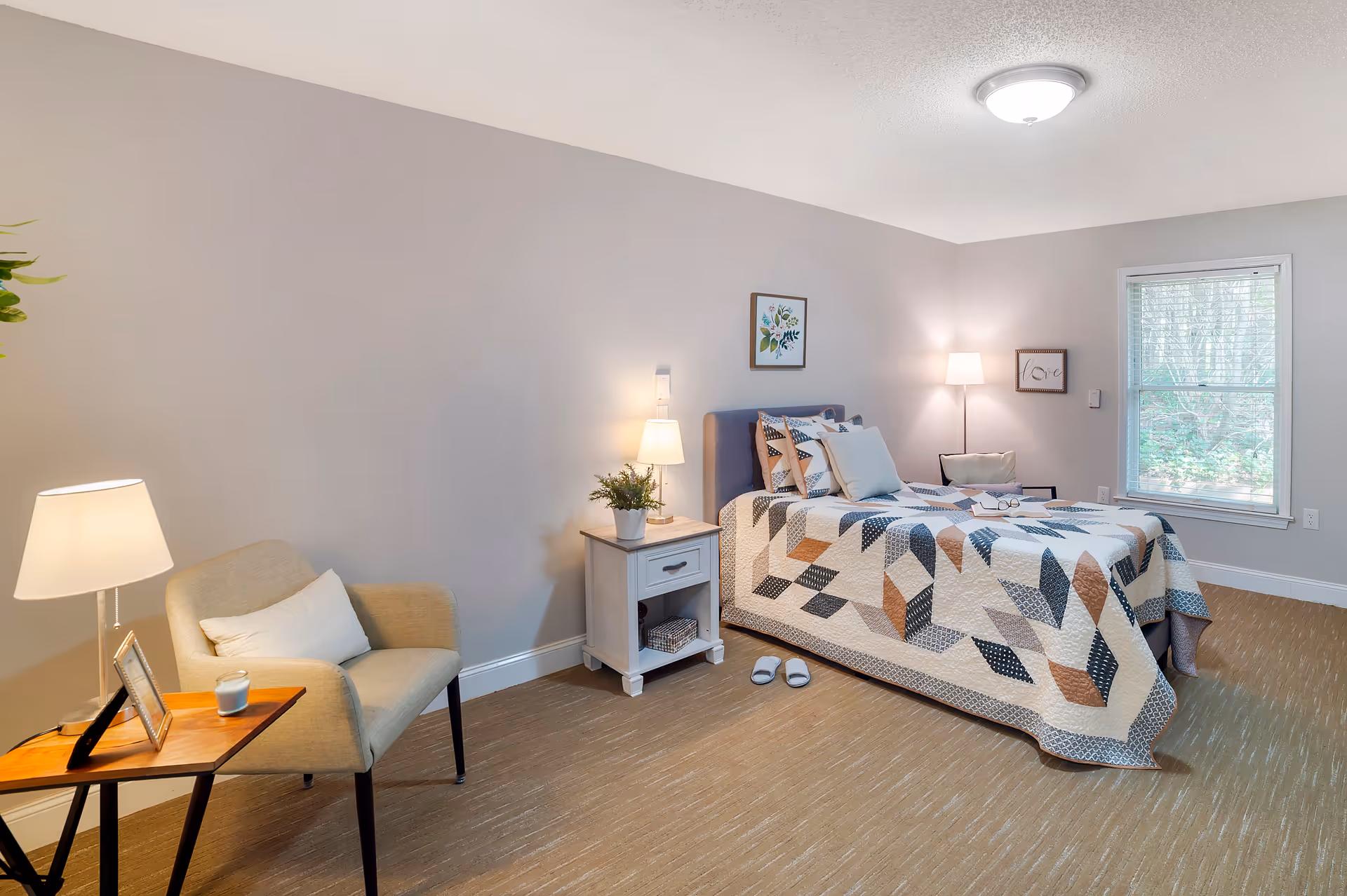 A cozy bedroom with a single bed covered in a patterned quilt, a nightstand with a lamp and a small plant, a beige armchair with a pillow next to a wooden side table holding a lamp and a candle, a window with blinds letting in natural light, and two framed pictures on the light gray walls.