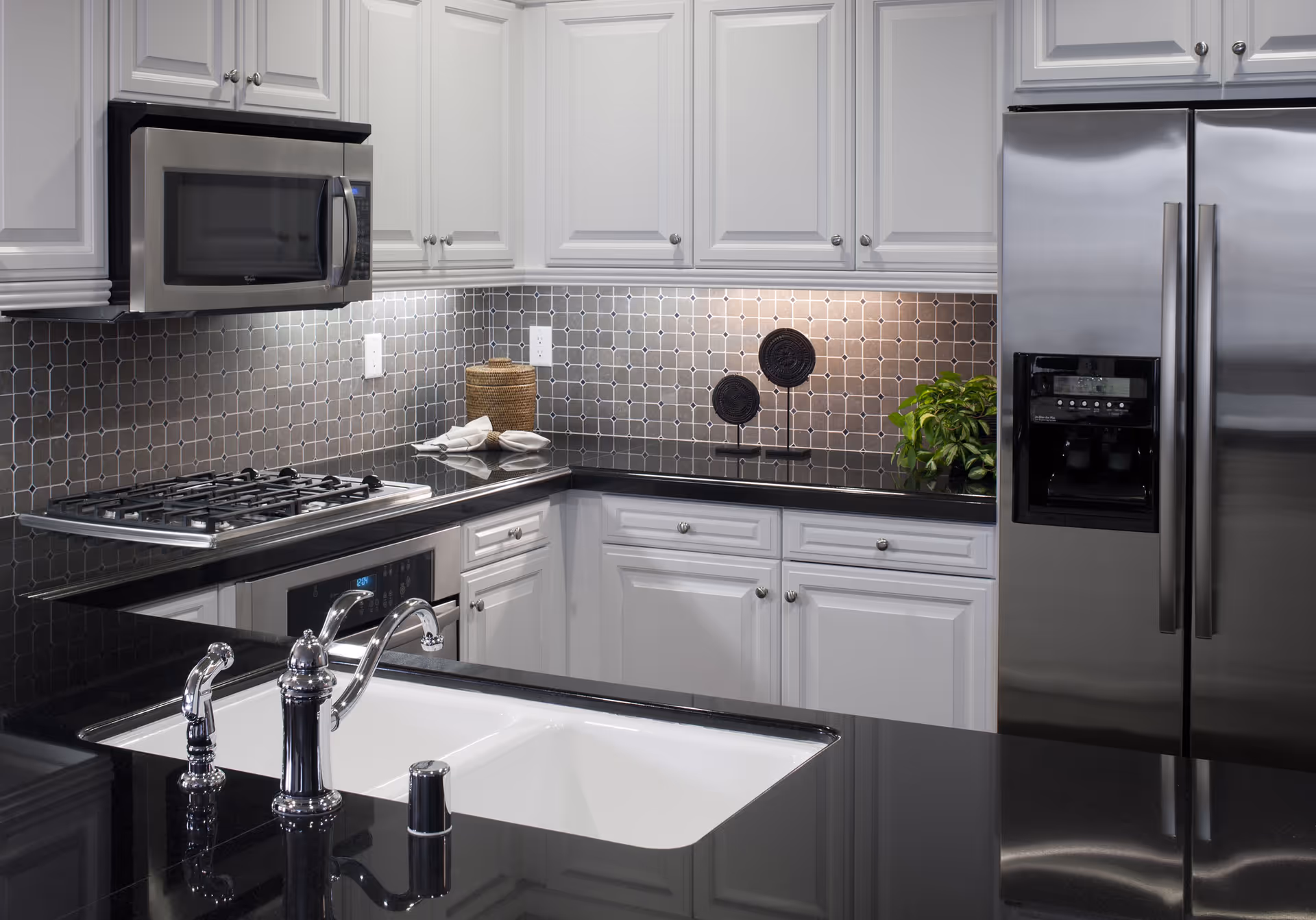 Modern kitchen with white cabinets, black countertops, a stainless steel microwave, gas stove, oven, and refrigerator. The backsplash features a gray tile pattern, and there is a white sink with a chrome faucet in the foreground. Decorative items and a small green plant are on the countertop.