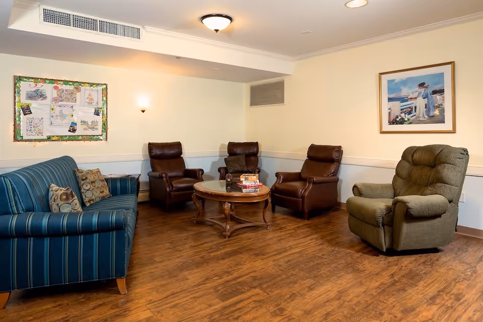 A cozy sitting area in a memory care facility with a striped blue sofa, three brown leather armchairs, and one green recliner arranged around a round wooden coffee table with books on it. The room has wooden flooring, cream-colored walls, a bulletin board with papers and decorations, and a framed painting of two women by the sea.