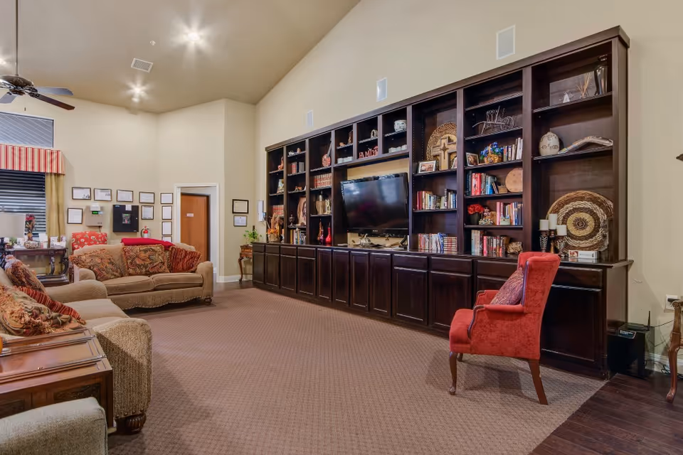 A cozy living room area in a senior living facility featuring two beige sofas with patterned cushions, a red armchair, and a large dark wooden built-in bookshelf with a mounted flat-screen TV. The bookshelf is decorated with books, framed photos, and various decorative items. The room has beige walls, carpeted flooring, and ceiling lights.