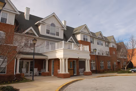 Front entrance of a multi-story brick and siding senior living building with a covered portico, balcony, and rocking chairs.