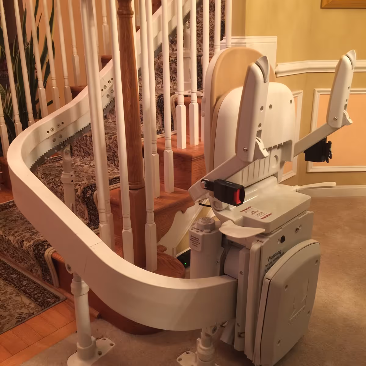 A stairlift installed on a carpeted staircase with wooden steps and white railings inside a home or facility. The stairlift chair is folded up, showing its white and beige components and safety features.