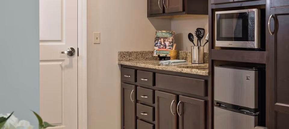 A kitchenette area with dark wood cabinets, a granite countertop, a microwave, and a small refrigerator. Kitchen utensils are stored in a container on the counter, and a cookbook is displayed on a stand. A closed white door is visible to the left.