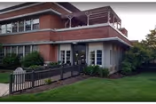 Front entrance of a two-story brick senior living building with a covered porch, balcony, ramp, and surrounding lawn.
