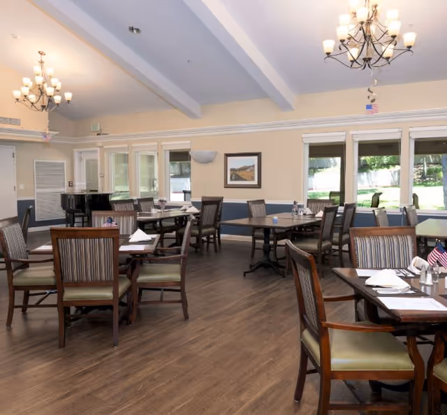 A bright senior living dining room with multiple wooden tables and chairs, chandeliers, large windows, and a piano in the back.