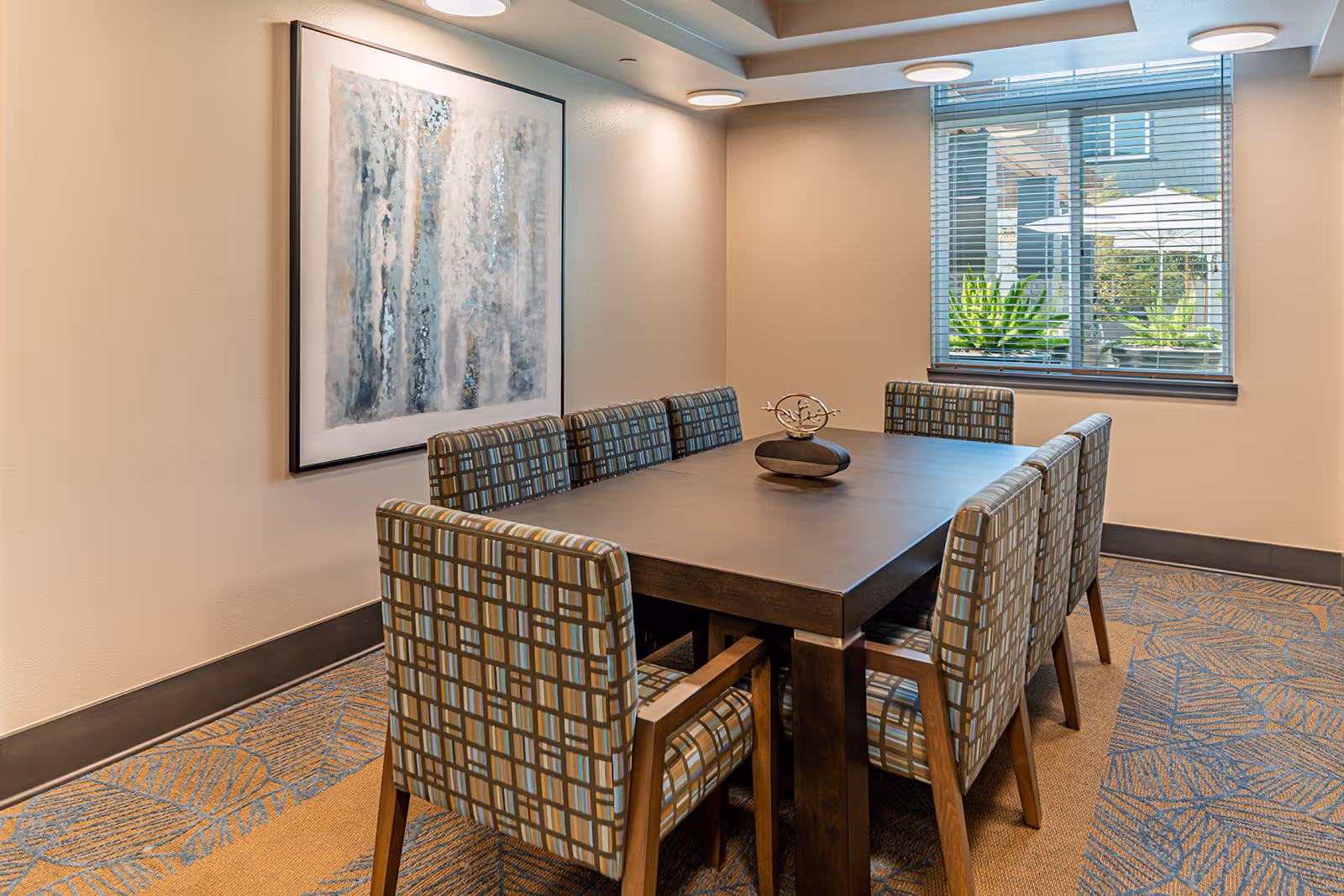 A modern dining room with a rectangular dark wooden table surrounded by eight patterned upholstered chairs. A decorative centerpiece sits on the table. The room has beige walls, a large abstract painting on one wall, and a window with blinds showing outdoor greenery and umbrellas. The floor is carpeted with a blue and tan leaf pattern.