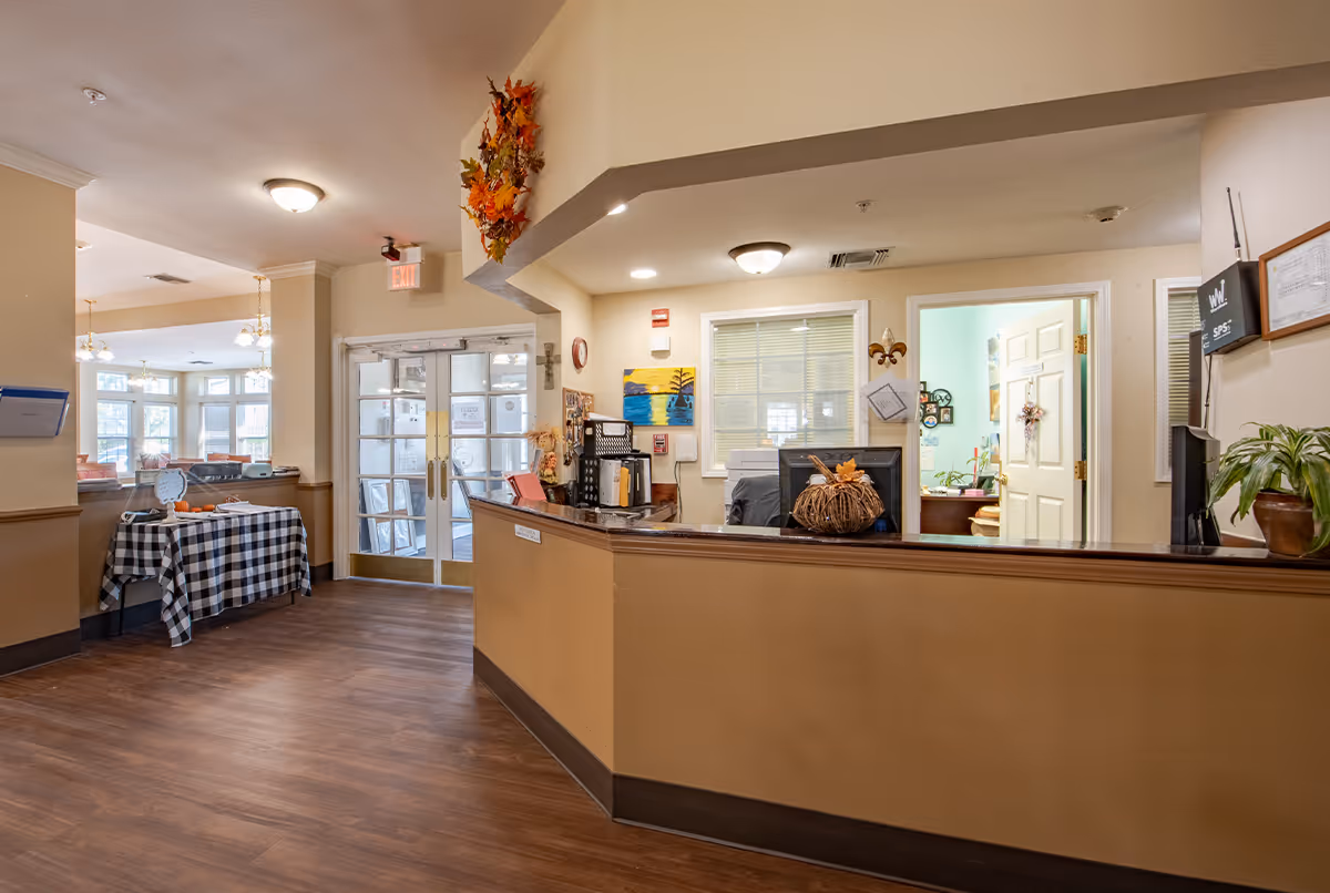 Reception area of a senior living facility with a wooden front desk decorated with a small pumpkin and fall leaves. Behind the desk are office supplies, a computer monitor, and a window looking into an office. To the left, double glass doors lead outside, and a table covered with a black and white checkered cloth holds some items. The area is well-lit with ceiling lights and has wood flooring.