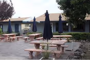 Outdoor patio area with multiple stone picnic tables and benches, each with a closed dark blue umbrella. The patio is paved with bricks and surrounded by greenery including bushes and trees. A single-story building with a beige roof and light-colored walls is visible in the background.