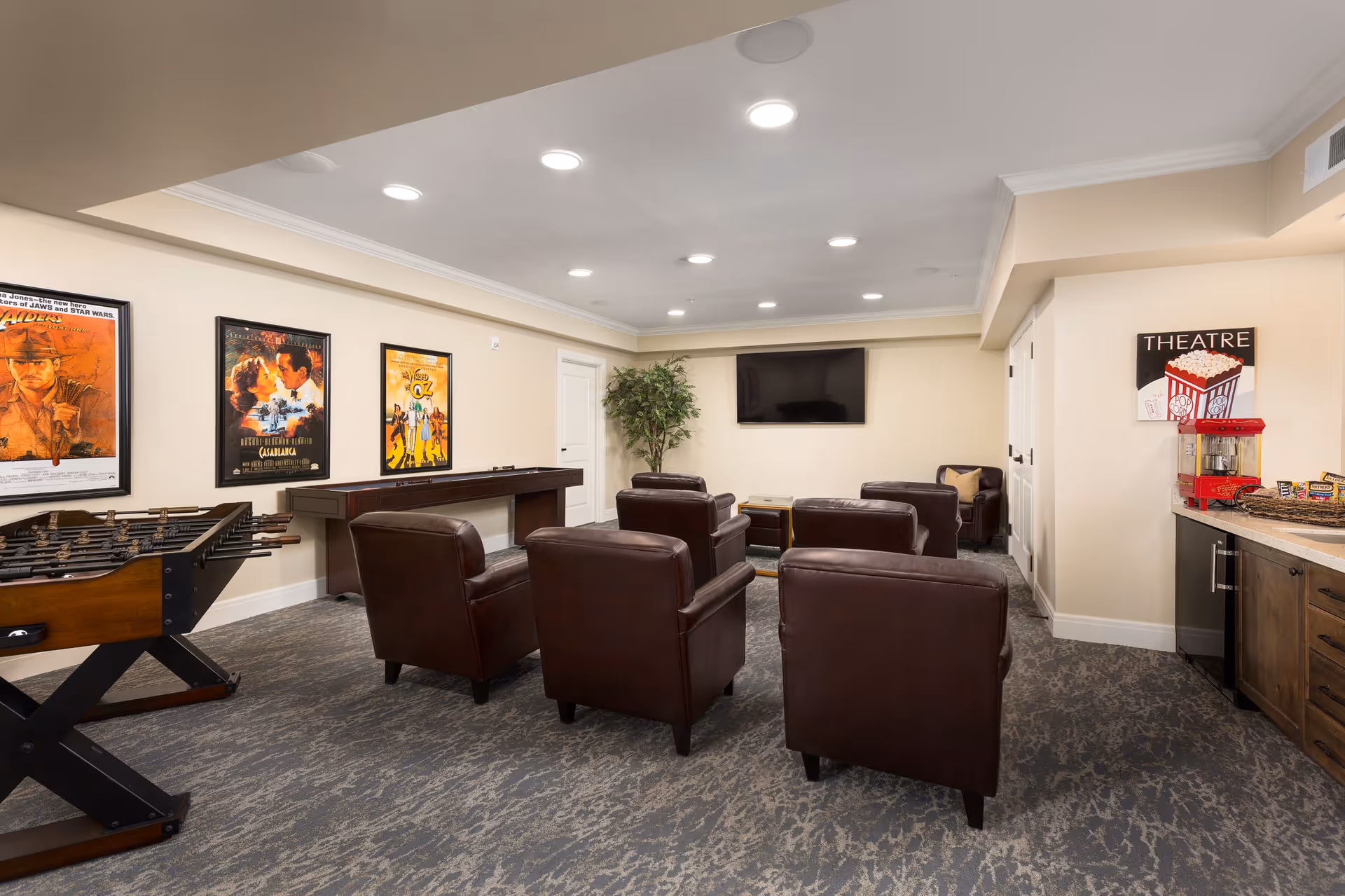 A cozy entertainment room with six brown leather armchairs arranged in two rows facing a wall-mounted flat-screen TV. The room features a foosball table on the left, movie posters on the wall, a shuffleboard table, a popcorn machine on a countertop to the right, and a potted plant in the corner. The carpet has a patterned design and the walls are painted a light beige color.