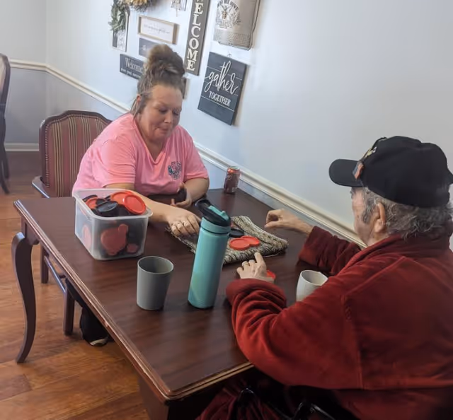 Two people sitting at a wooden table playing a game with red circular pieces. One person is wearing a pink shirt and has their hair in a bun, while the other person is wearing a red jacket and a black cap. The table has a plastic container with more game pieces, two cups, and a water bottle. The wall behind them has decorative signs with welcoming messages.
