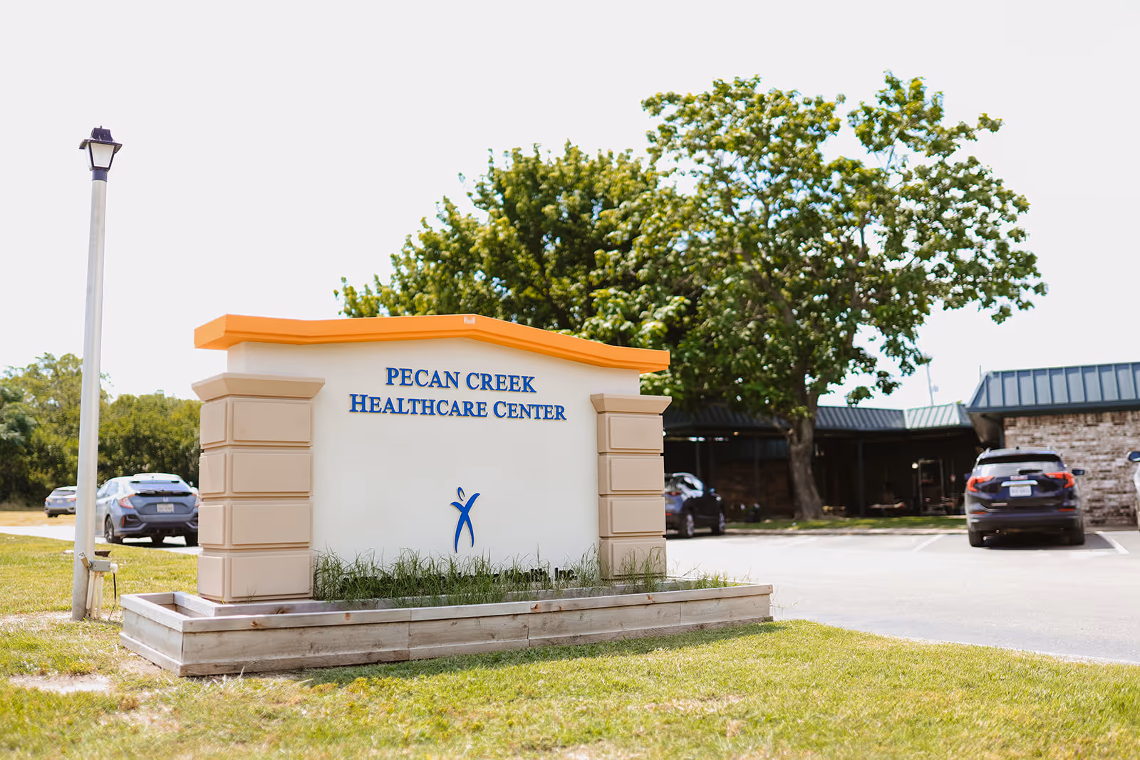 Outdoor sign reading "Pecan Creek Healthcare Center" in front of the facility with cars, a tree, and the building visible behind it.