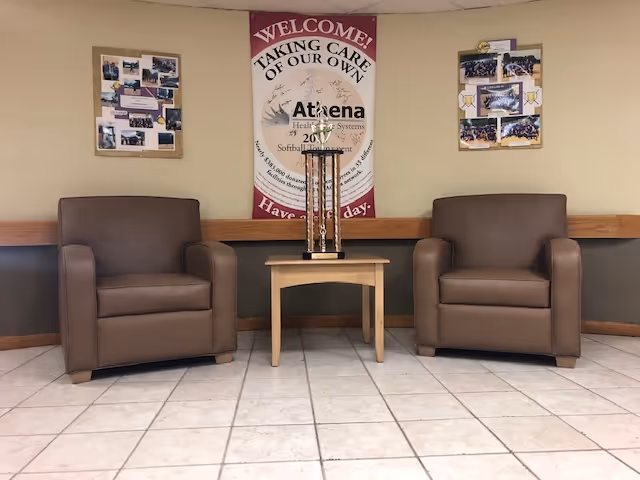 Two brown leather armchairs with a small wooden table between them, holding a tall trophy. Behind the chairs on the wall is a banner that reads 'WELCOME! TAKING CARE OF OUR OWN Athena Health Systems 2017 Softball Tournament Have a nice day.' There are also two bulletin boards with photos on either side of the banner. The floor is tiled with light-colored tiles.