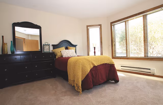 A bright bedroom with a single bed covered in a burgundy bedspread and a mustard yellow throw blanket. The bed has multiple pillows and a dark wooden headboard. Next to the bed is a black dresser with a large mirror, decorative vases, and a clock. The room has beige carpet and large windows with wooden blinds letting in natural light.