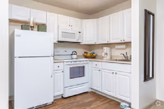 A clean kitchen with white cabinets, a white refrigerator, a white stove with an oven, a microwave above the stove, a coffee maker on the countertop, a bowl of fruit, and wooden flooring.