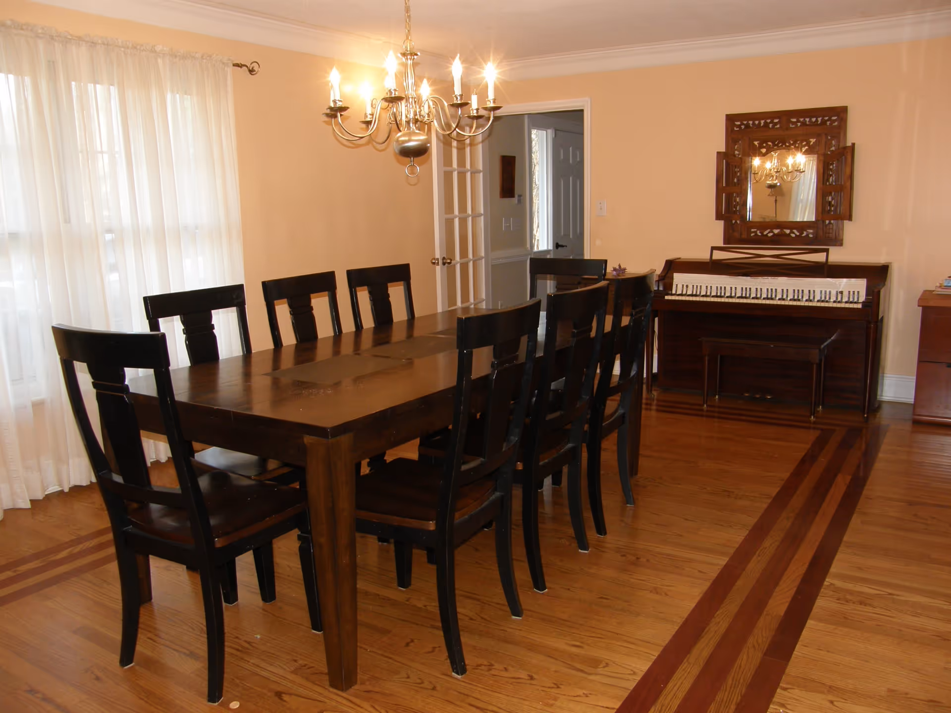 A dining room with a long wooden table surrounded by eight dark wooden chairs. A chandelier with lit bulbs hangs above the table. There is a piano against the far wall with a decorative wooden framed mirror above it. The room has light-colored walls and wooden flooring with a decorative inlay. Sheer white curtains cover the windows on the left side.