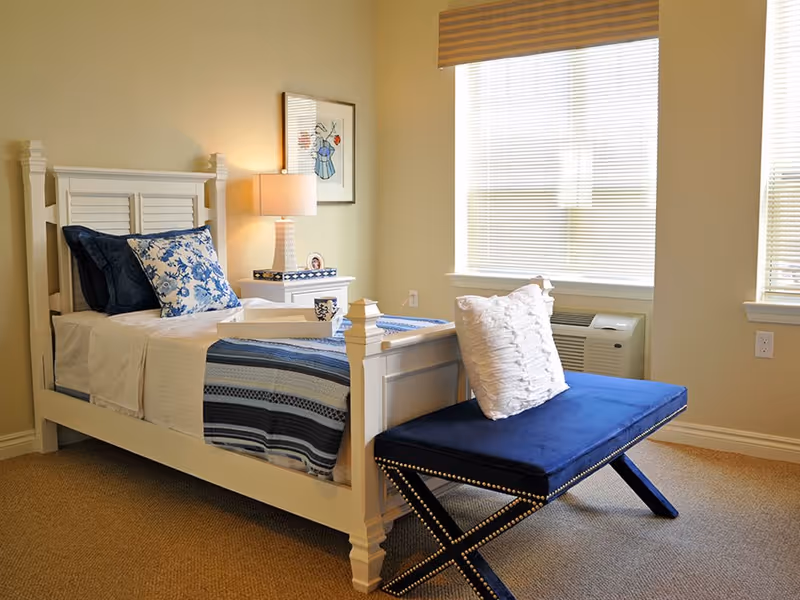 A neatly furnished bedroom with a white wooden bed, blue patterned bedding, a bedside table and lamp, and a blue upholstered bench by a window.